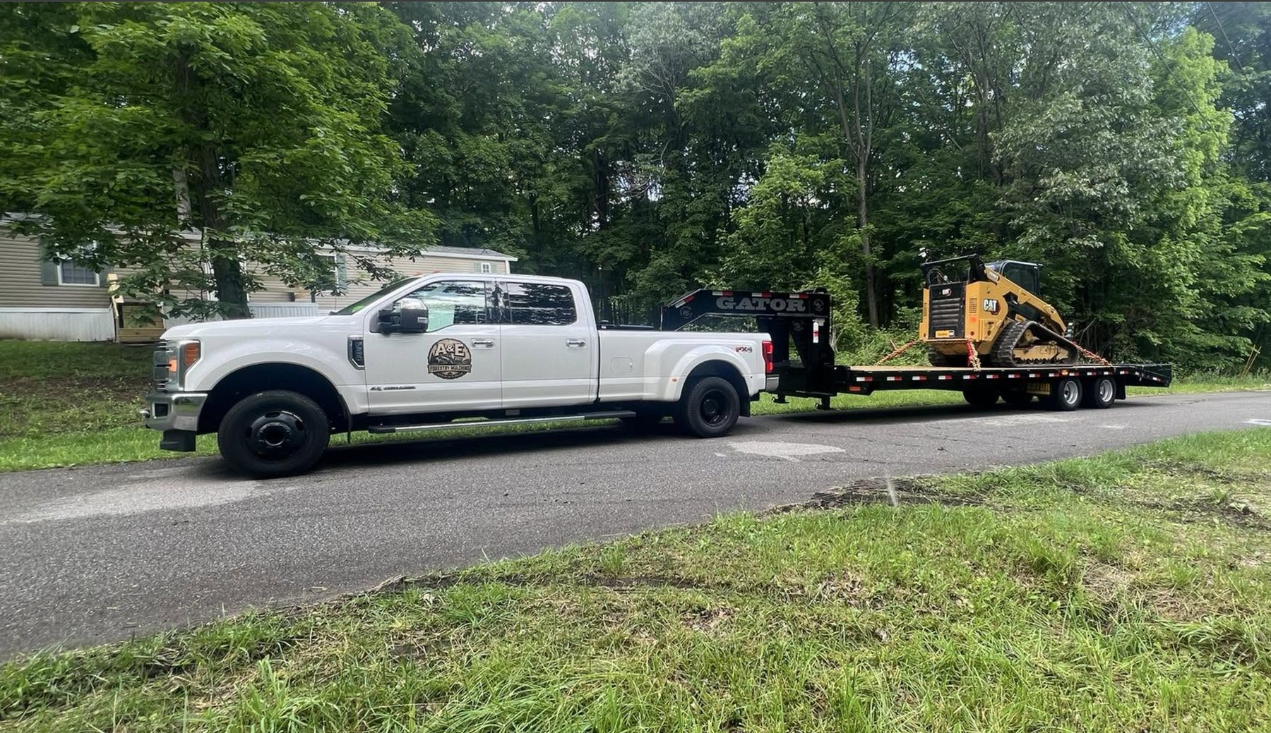 A&E Forestry Mulching's white dually with the company logo on the door, towing a gooseneck trailer with a tracked CAT skid steer and mulching head loaded on it.